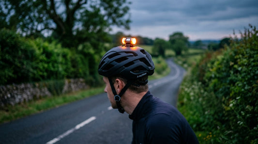 A road cycling helmet with a compact LED helmet light mounted on top, glowing at dusk on a UK country lane, hedgerow blurred in the background